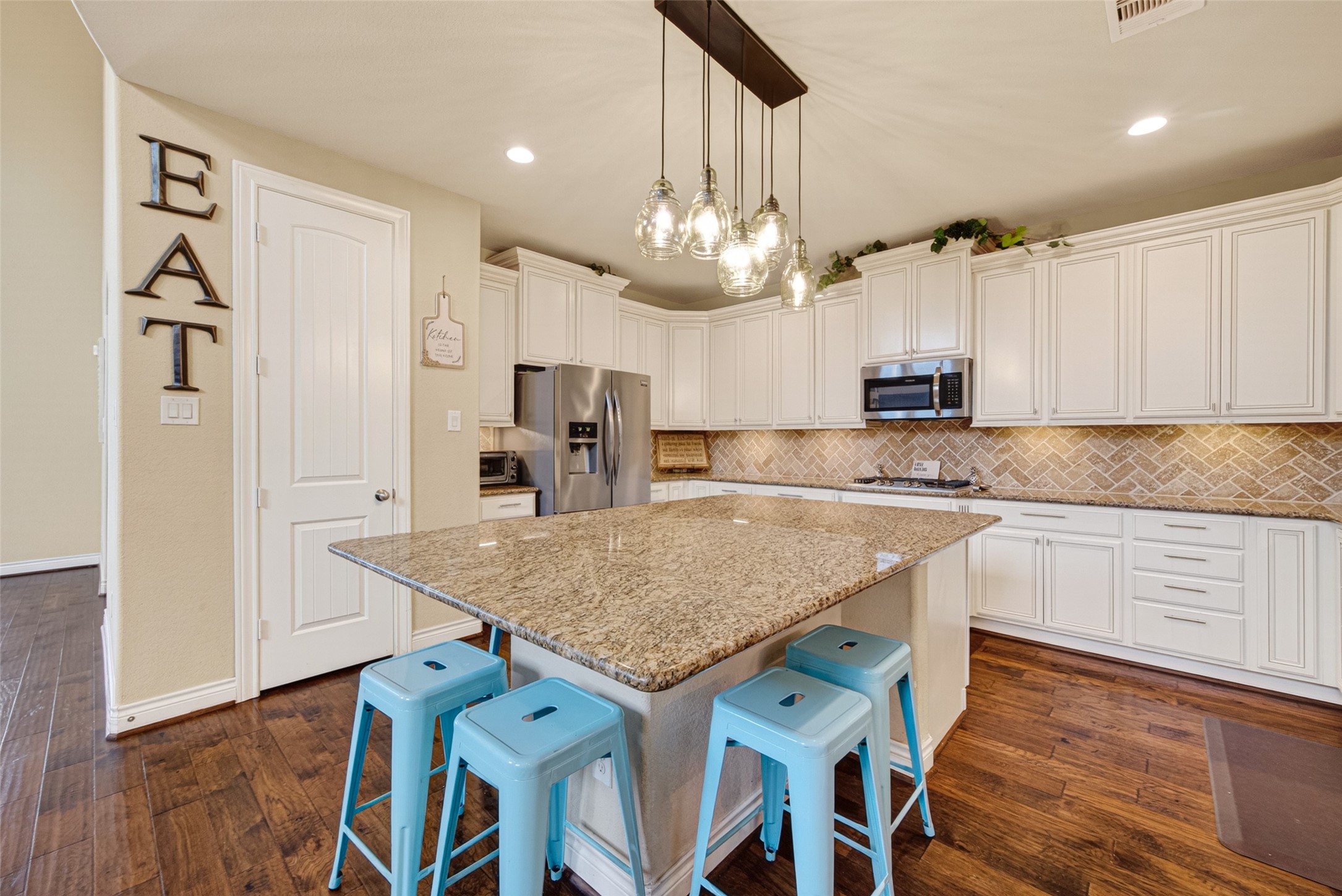 25611 Ellis Ridge Court Katy, TX 77494 - Photo 12 of 49 This spacious kitchen features a large granite island with seating, stylish pendant lighting, and ample white cabinetry. The stainless steel appliances and herringbone backsplash add a modern touch, while the warm wood floors create a cozy atmosphere.
