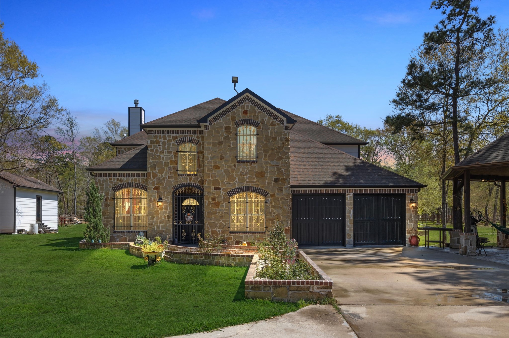 This photo showcases a charming stone-accented two-story home with a three-car garage. It features a spacious driveway, lush green lawn, and is surrounded by mature trees, offering a serene and inviting atmosphere.