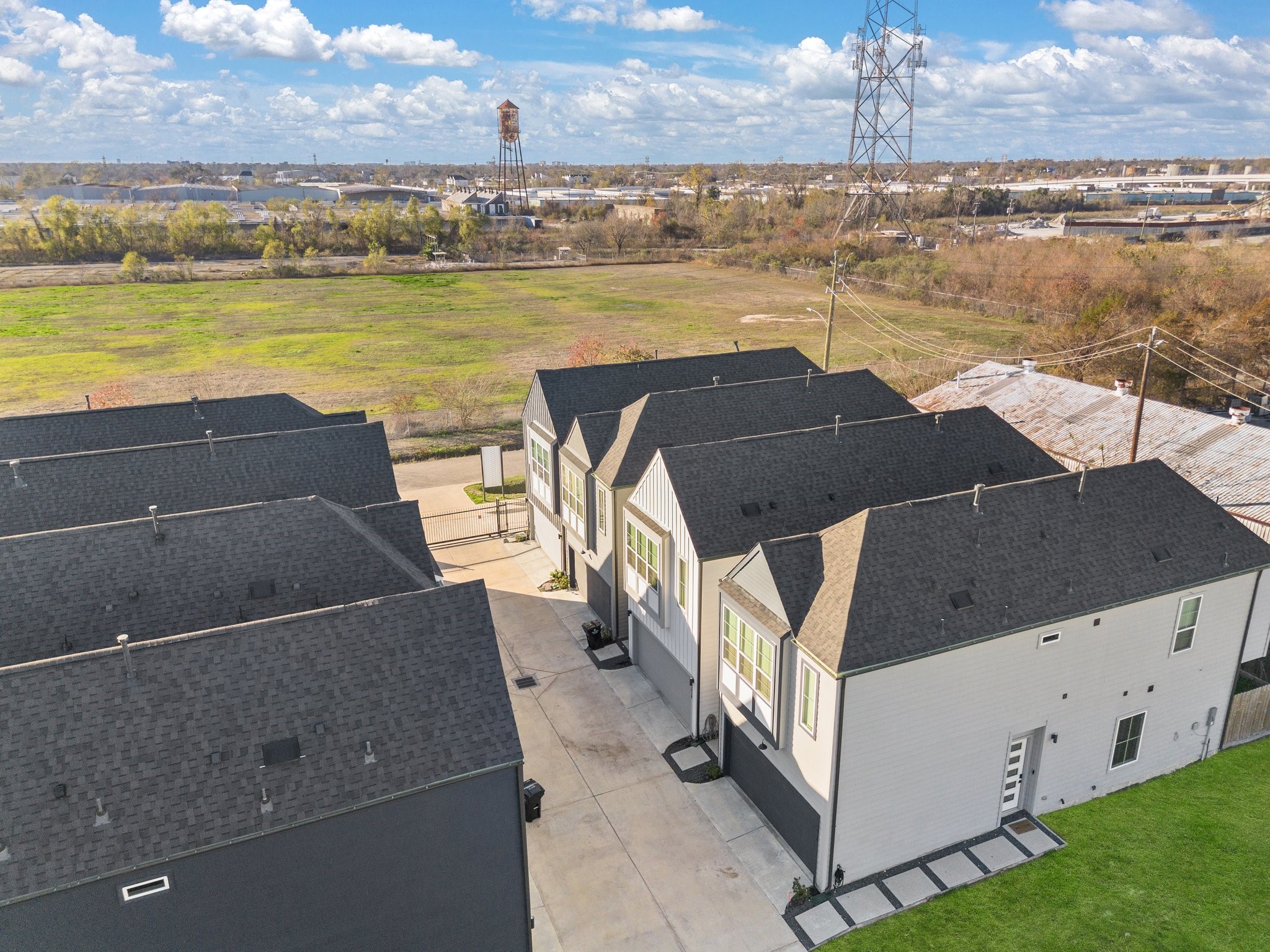 2928 Stevens Street Houston, TX 77026 - Photo 24 of 30 Modern townhomes with dark roofs are lined up along a concrete driveway. The area is open, with green fields and distant industrial structures visible. The location offers a spacious, suburban feel.