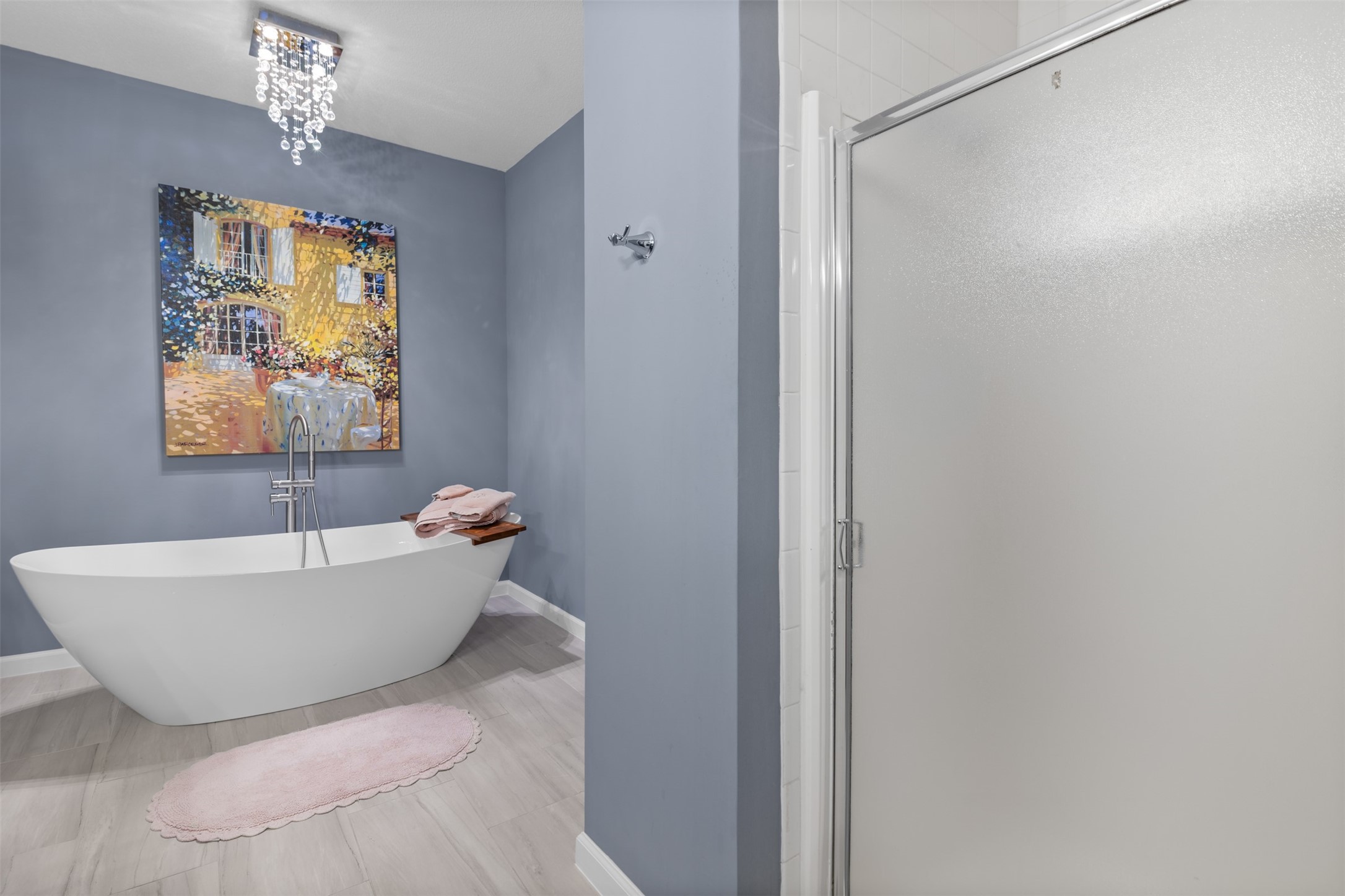 6000 Reims Road, Unit 2307 Houston, TX 77036 - Photo 15 of 26 This bathroom features a modern freestanding bathtub with elegant fixtures, a stylish blue-gray wall, and a framed artwork. A sleek glass shower enclosure is adjacent, and a decorative light fixture adds a touch of luxury.