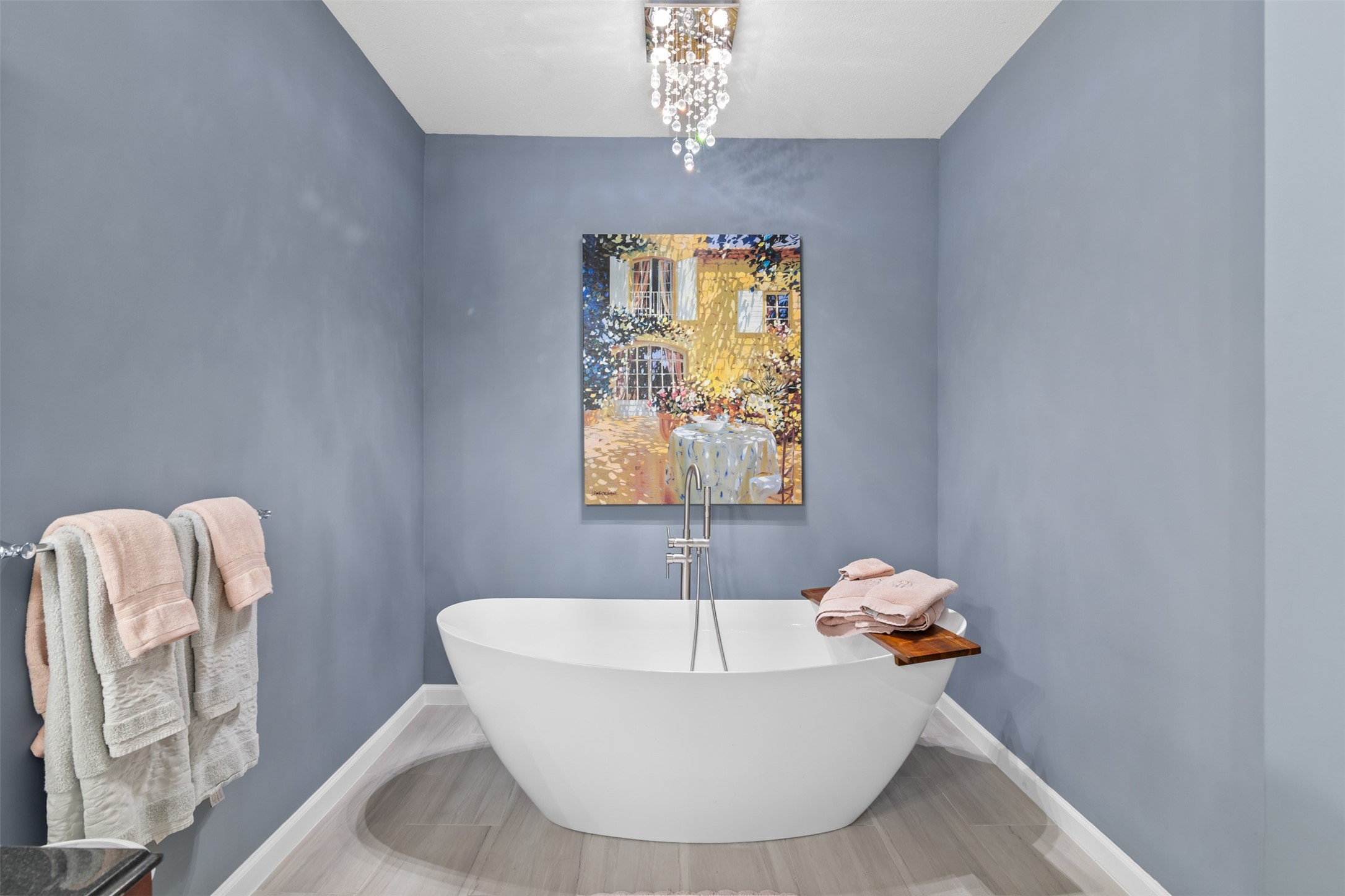 6000 Reims Road, Unit 2307 Houston, TX 77036 - Photo 16 of 26 This modern bathroom features a freestanding tub against a soft blue wall, accented by a vibrant painting. A sleek chandelier adds elegance, while neatly arranged towels and a wooden bath tray enhance functionality and style.