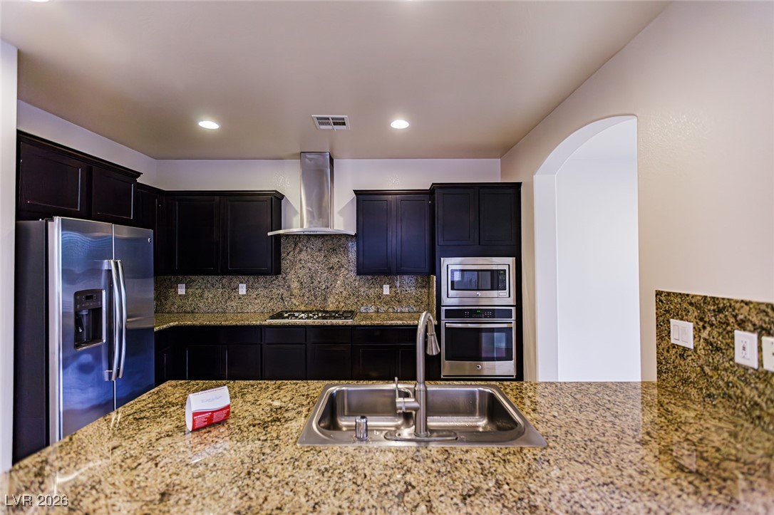 11009 Ketchikan Street Las Vegas, NV 89179 - Photo 10 of 59 Kitchen with decorative backsplash, appliances with stainless steel finishes, wall chimney range hood, light stone countertops, and arched walkways