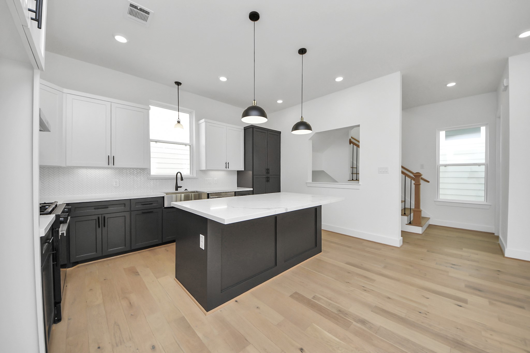 723 Marcella Street, Unit B Houston, TX 77091 - Photo 3 of 22 This modern kitchen features sleek black and white cabinetry, a central island large island with a white countertop, and stylish pendant lighting. It has light wood flooring and an open layout, with stairs visible in the background.