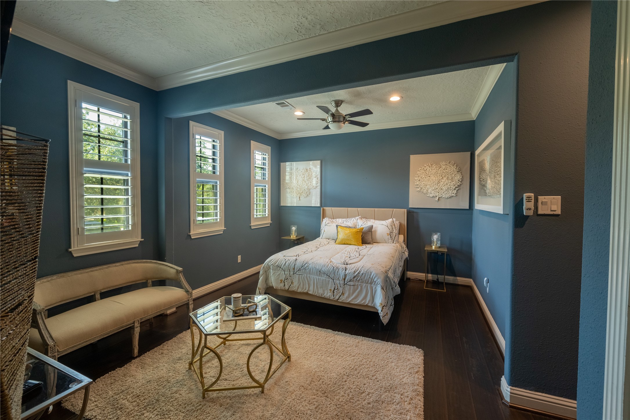 4002 Silver Ridge Boulevard Missouri City, TX 77459 - Photo 32 of 50 This is a spacious (Guest Quarters) bedroom featuring dark wood flooring, deep blue walls with a tray ceiling, and several windows providing natural light. The room is furnished with a comfortable bed, a bench, a small table, and two wall art pieces. A ceiling fan offers additional comfort.
