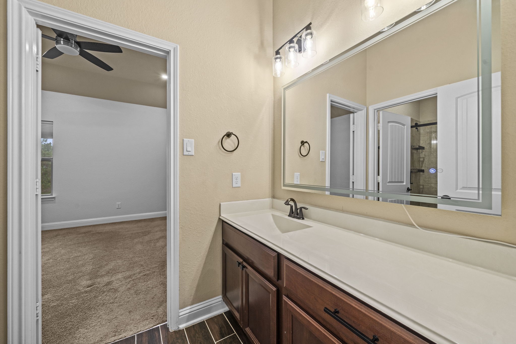 14215 Spindle Arbor Road Cypress, TX 77429 - Photo 27 of 47 This photo showcases a bathroom with a large mirror and a single-sink vanity featuring dark wood cabinetry. The space is well-lit with modern fixtures, and there is a glimpse into an adjacent carpeted room with a ceiling fan.