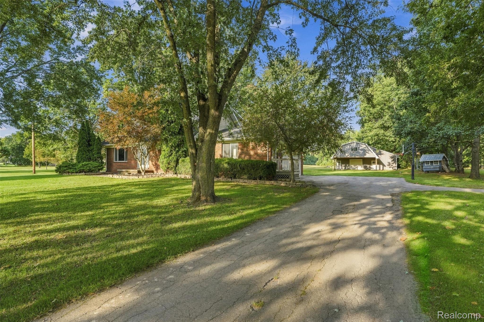 Brick exterior home with a paved driveway, expansive green lawn, mature trees, and an additional detached structure