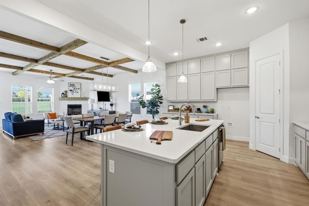 4017 Eagles Bluff Road Midlothian, TX 76065 - Photo 6 of 38 Open-concept kitchen and living area featuring a large center island with a stainless steel sink, light gray cabinetry, wood-finish flooring, exposed ceiling beams, and a fireplace with a white mantel