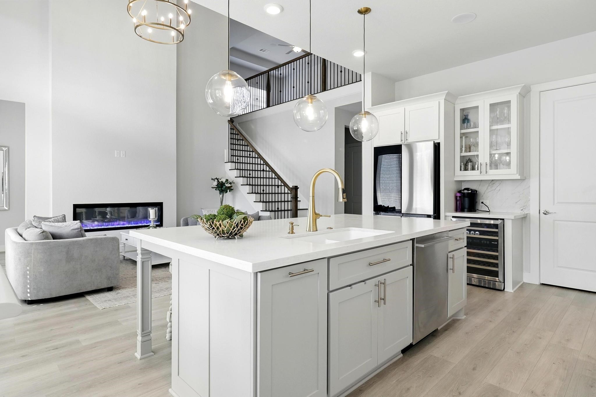 26923 Arethusa Court Spring, TX 77389 - Photo 11 of 44 This modern kitchen features sleek white cabinetry, a spacious island with a gold-tone faucet, and pendant lighting. The open layout connects to a living area with a cozy fireplace and staircase, offering a bright and inviting space.