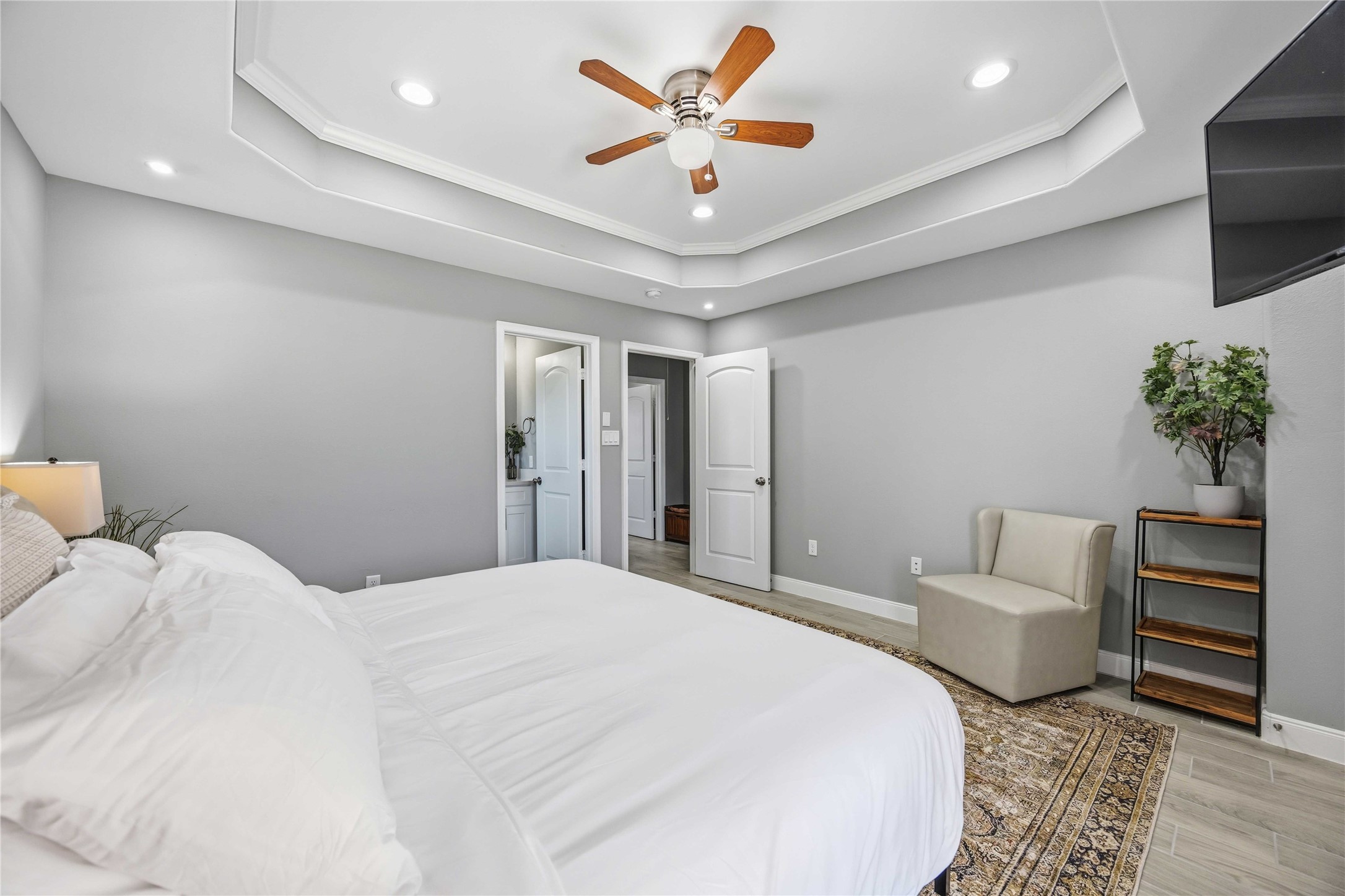 4903 Mallow Street, Unit A Houston, TX 77033 - Photo 7 of 10 This master bedroom features a clean, contemporary design with a neutral color palette of cool grays and crisp whites. The space feels open and airy. The standout feature is a large double recessed (tray) ceiling with crown molding. It includes a central five-blade wood ceiling fan with an integrated light fixture, supplemented by several recessed LED "can" lights for even illumination.