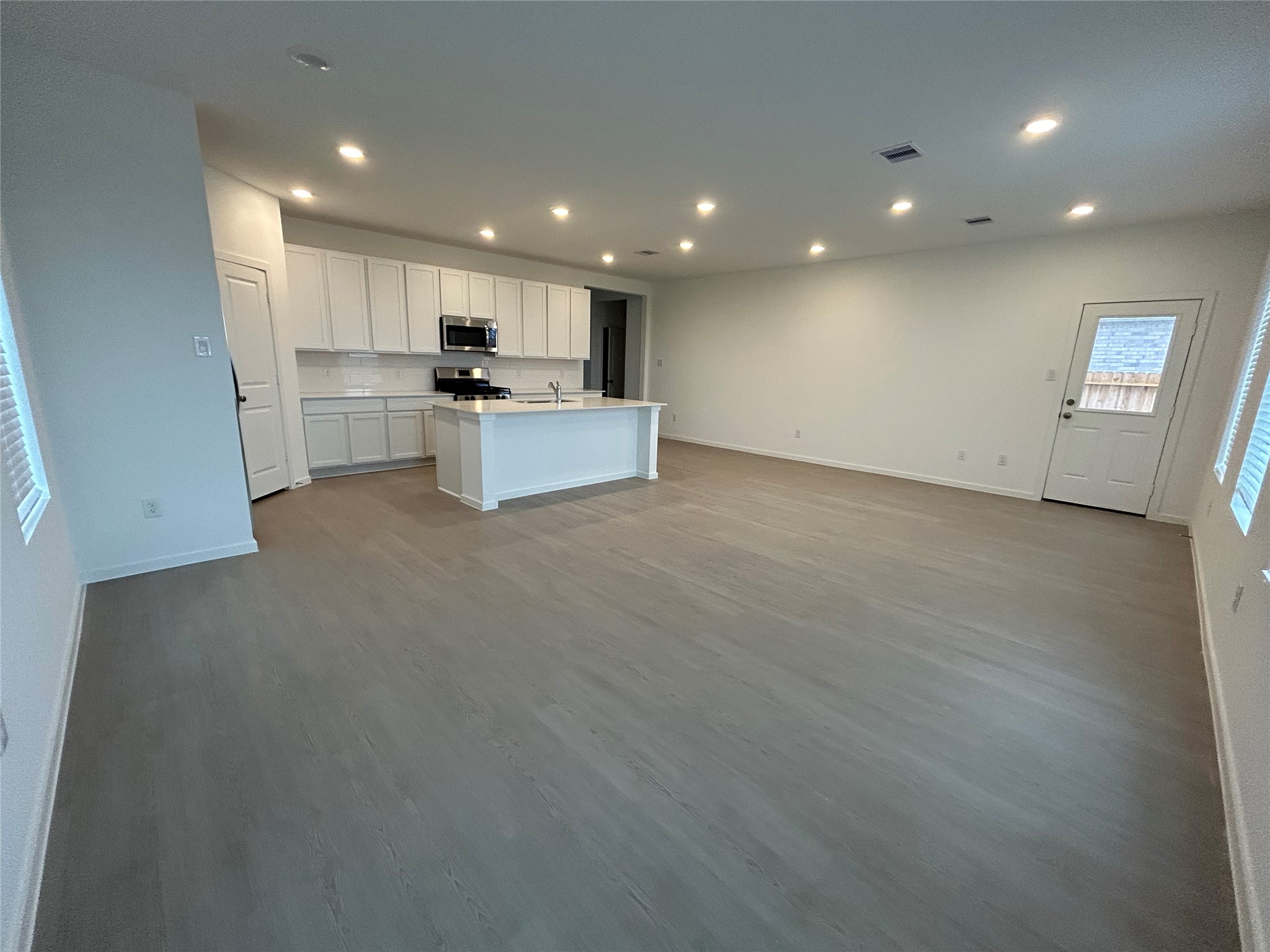 26419 Virginia Wild Rye Lane Katy, TX 77493 - Photo 11 of 31 This photo shows a spacious, open-concept kitchen and living area with modern finishes. It features sleek white cabinetry, a large island with a sink, recessed lighting, and ample natural light from multiple windows. The flooring is a contemporary wood-like laminate, and the room has a clean, neutral color scheme.