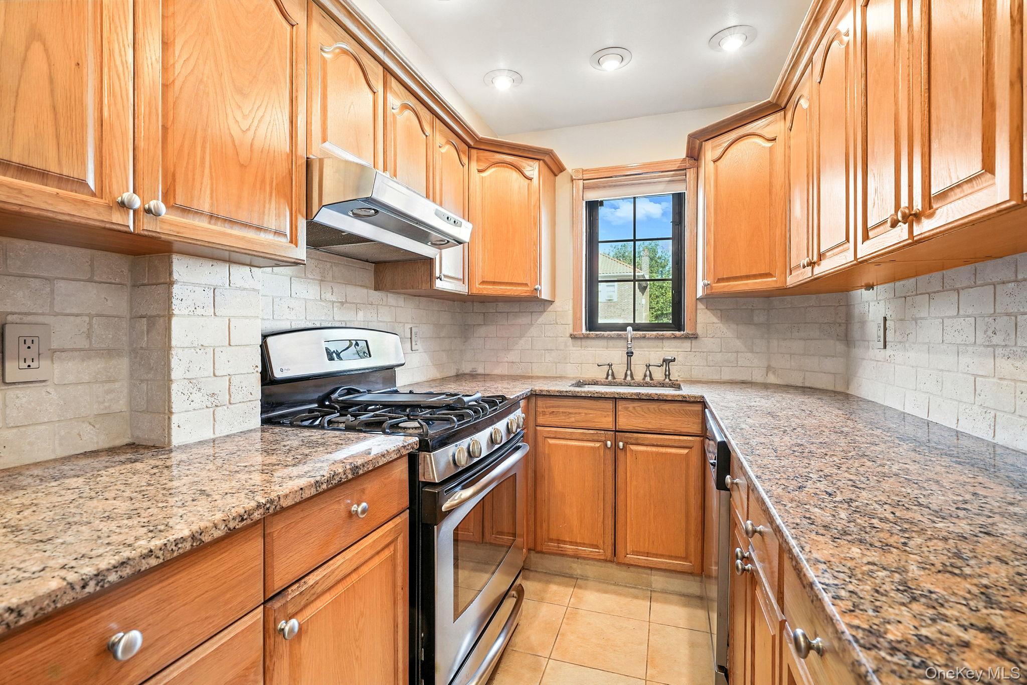 1245 Rhinelander Avenue Bronx, NY 10461 - Photo 12 of 34 Kitchen featuring appliances with stainless steel finishes, under cabinet range hood, light stone counters, light tile patterned floors, and brown cabinetry