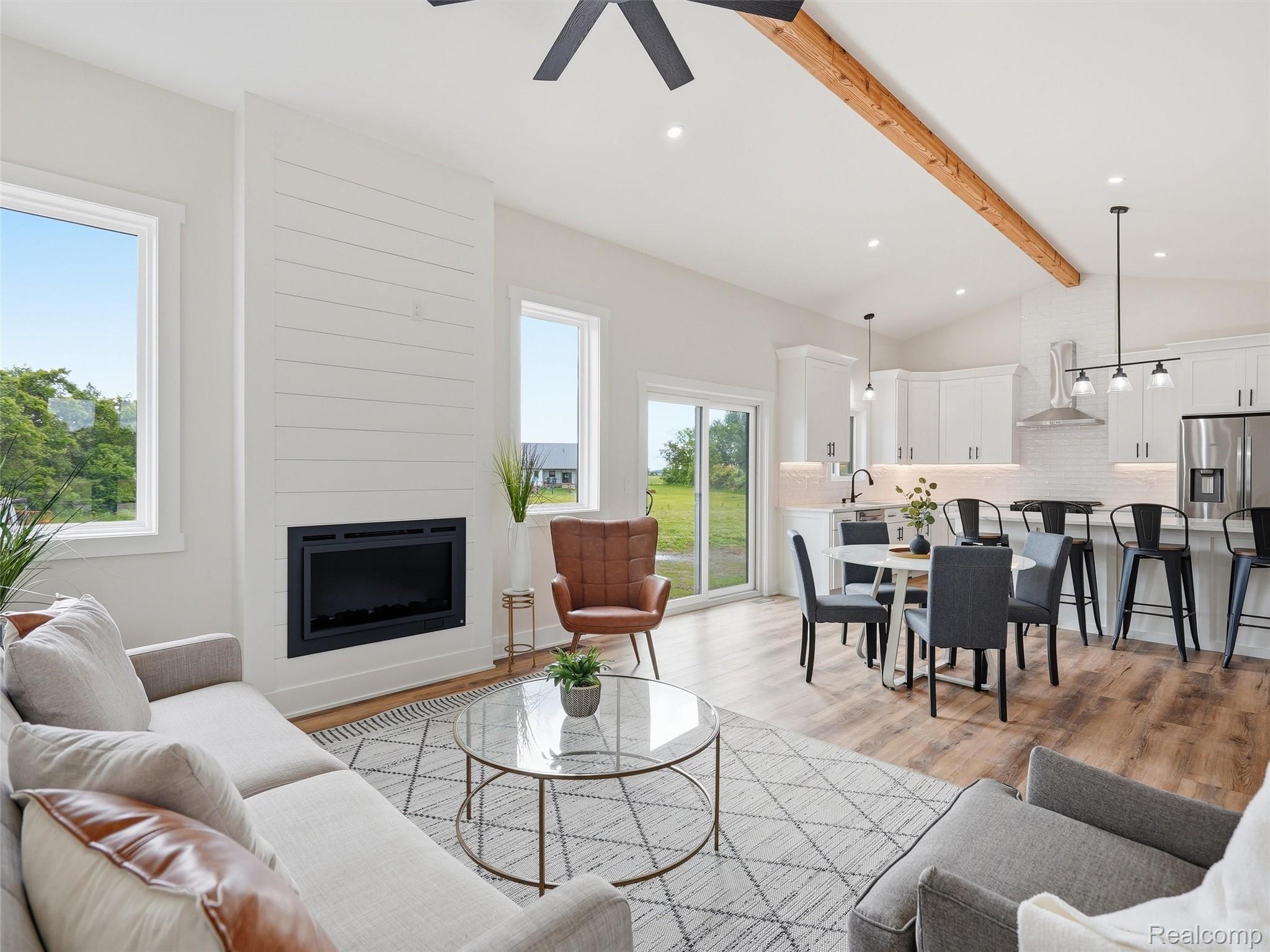 77850 Angling Road Richmond, MI 48062 - Photo 11 of 54 Open-concept living space featuring vaulted ceilings with exposed wood beams, a modern fireplace with shiplap surround, wood-finish flooring, and white kitchen cabinetry with stainless steel appliances