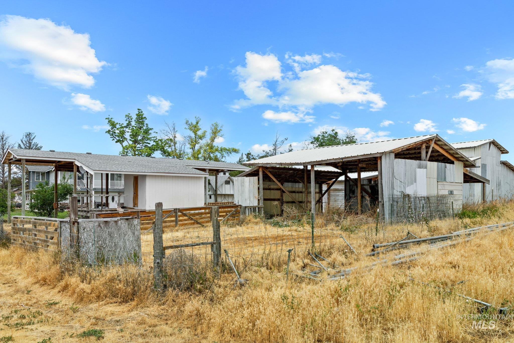 18338 Pride Lane Caldwell, ID 83607 - Photo 45 of 50 Property featuring a white-sided residence with an extended covered deck, alongside multiple outbuildings with metal roofs and exposed timber framing, all situated within an expansive dry grass landscape