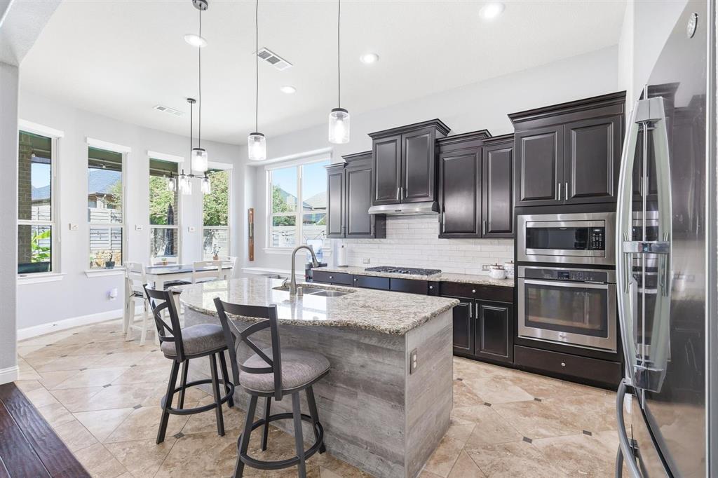 117 Birdcall Lane Argyle, TX 76226 - Photo 20 of 40 Kitchen featuring appliances with stainless steel finishes, light stone countertops, tasteful backsplash, a center island with sink, and a kitchen breakfast bar