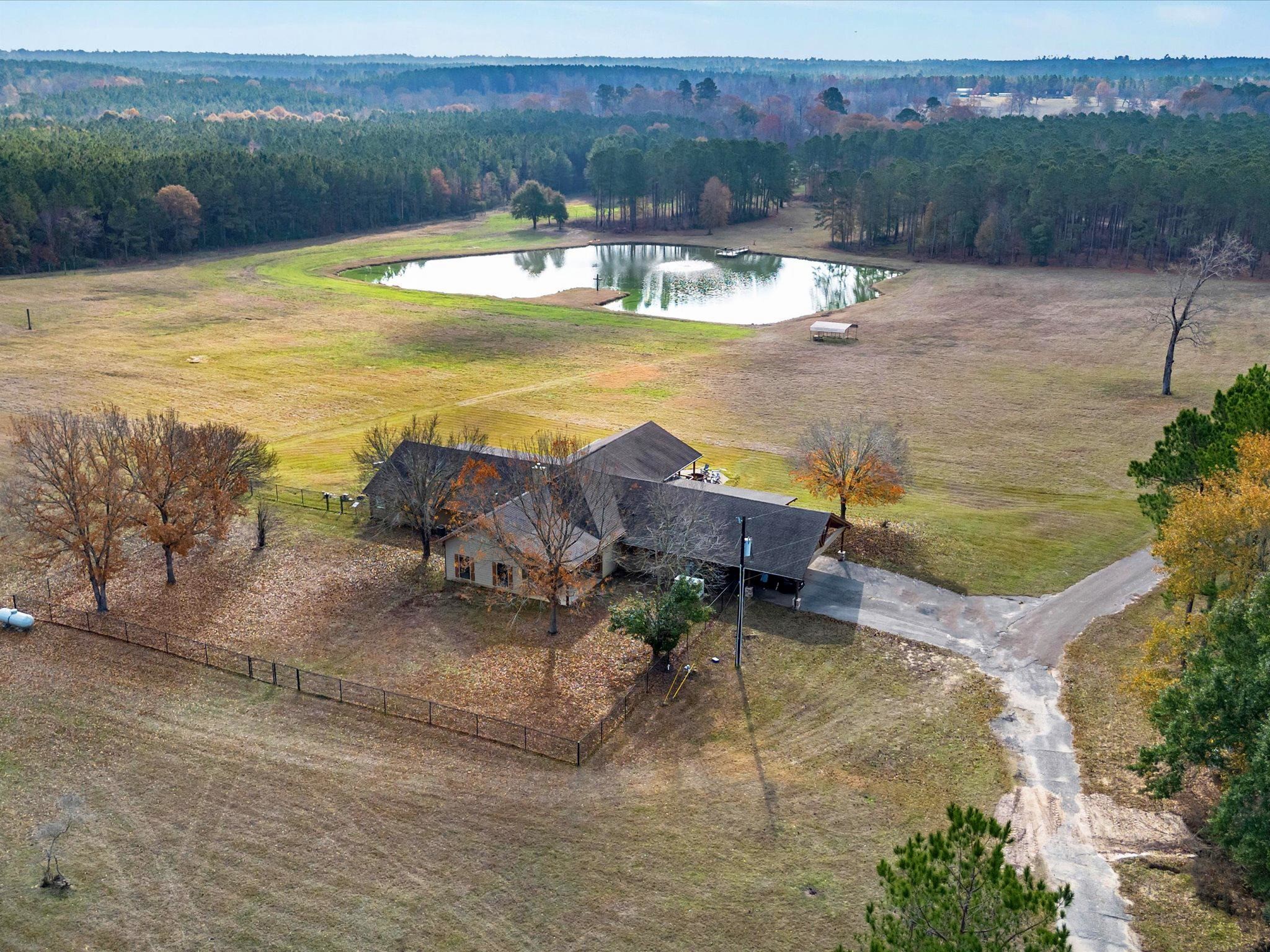This aerial view showcases a spacious rural property featuring a large, single-story home surrounded by open fields and mature trees. A serene pond is located nearby, providing a picturesque landscape. The property is accessed by a private driveway, offering a peaceful and secluded setting.