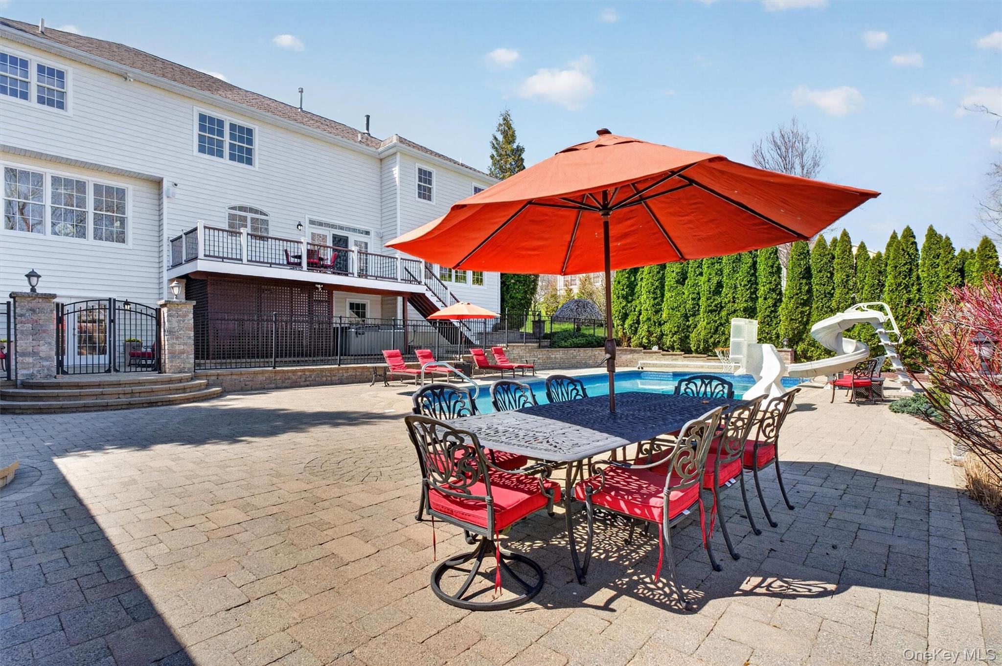 196 Country Club Road Hopewell Junction, NY 12533 - Photo 40 of 50 The lower patio is a true outdoor oasis, thoughtfully designed for both grand entertaining and everyday relaxation. Spanning an impressive 3,700 square feet, this custom Unilock paver patio is beautifully appointed with integrated lighting and anchored by a 7 Person PDC Spa & custom matching firepit—perfect for cozy evenings under the stars. The expansive layout easily accommodates full-scale outdoor dining and multiple seating areas, while the fire pit creates an inviting gathering space. A staircase provides seamless access to the upper deck, maximizing both flow and functionality. Enhancing the experience, a fully integrated outdoor sound system with 10 wired speakers—including pool rock, pool house, deck, under-deck, and fire pit placements—ensures the perfect ambiance throughout. An elegant black aluminum maintenance-free fence encloses and separates the in-ground pool area, offering both safety and a refined, unobtrusive design that complements the beauty of the surrounding landscape.