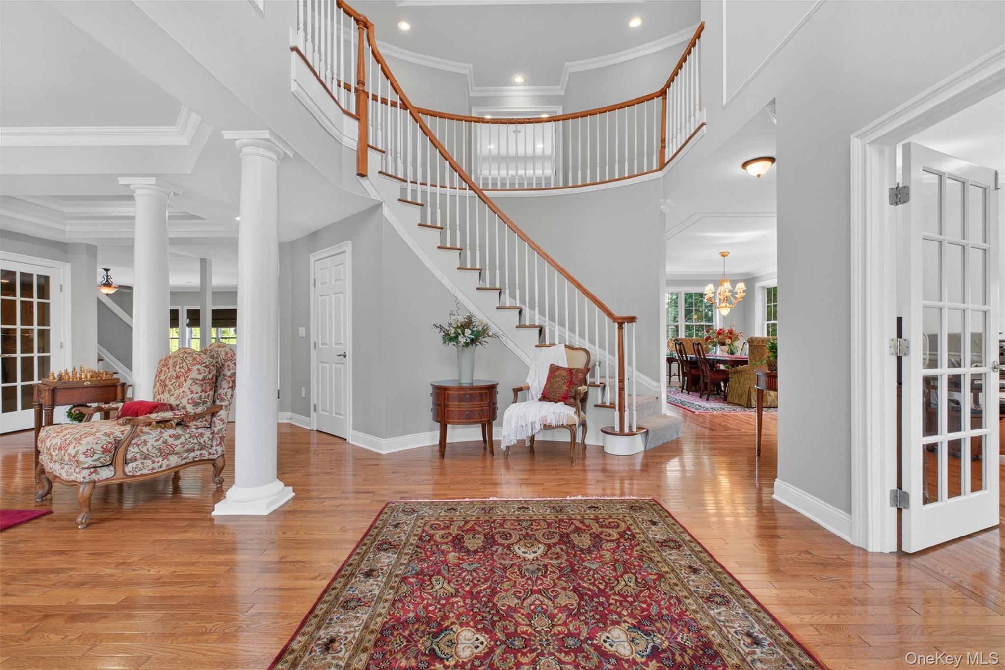 196 Country Club Road Hopewell Junction, NY 12533 - Photo 47 of 50 Stepping through the elegant glass front door, you are immediately welcomed into a breathtaking foyer of truly impressive scale—a masterclass in understated elegance—where volume, light, and architectural artistry take center stage. A sculptural, sweeping staircase of rich hardwood treads and matching hardwood railings curves gracefully upward, blending timeless grandeur with refined design. Affectionately referred to as a two story reception hall -The staircase flows effortlessly along the curved wall, its clean lines and elegant craftsmanship creating a striking yet harmonious focal point. From this grand vantage point, the openness of the home is immediately revealed—you can see into the formal living room accented by classic columns, the oversized executive office, the elegant dining room, and straight through to the rear of the home, offering a seamless and expansive floor plan. Above, a dazzling chandelier descends from the soaring two-story ceiling, its soft glow reflecting off polished hardwood floor