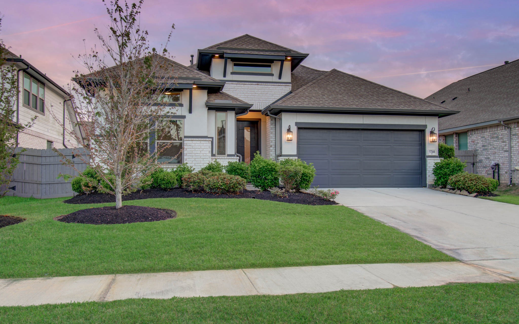 17344 Grand Canyon Road Conroe, TX 77302 - Photo 39 of 49 This photo showcases a modern, well-maintained two-story home with a gray exterior and a two-car garage. It features a landscaped front yard with a small tree and shrubbery, set against a beautiful sunset sky.