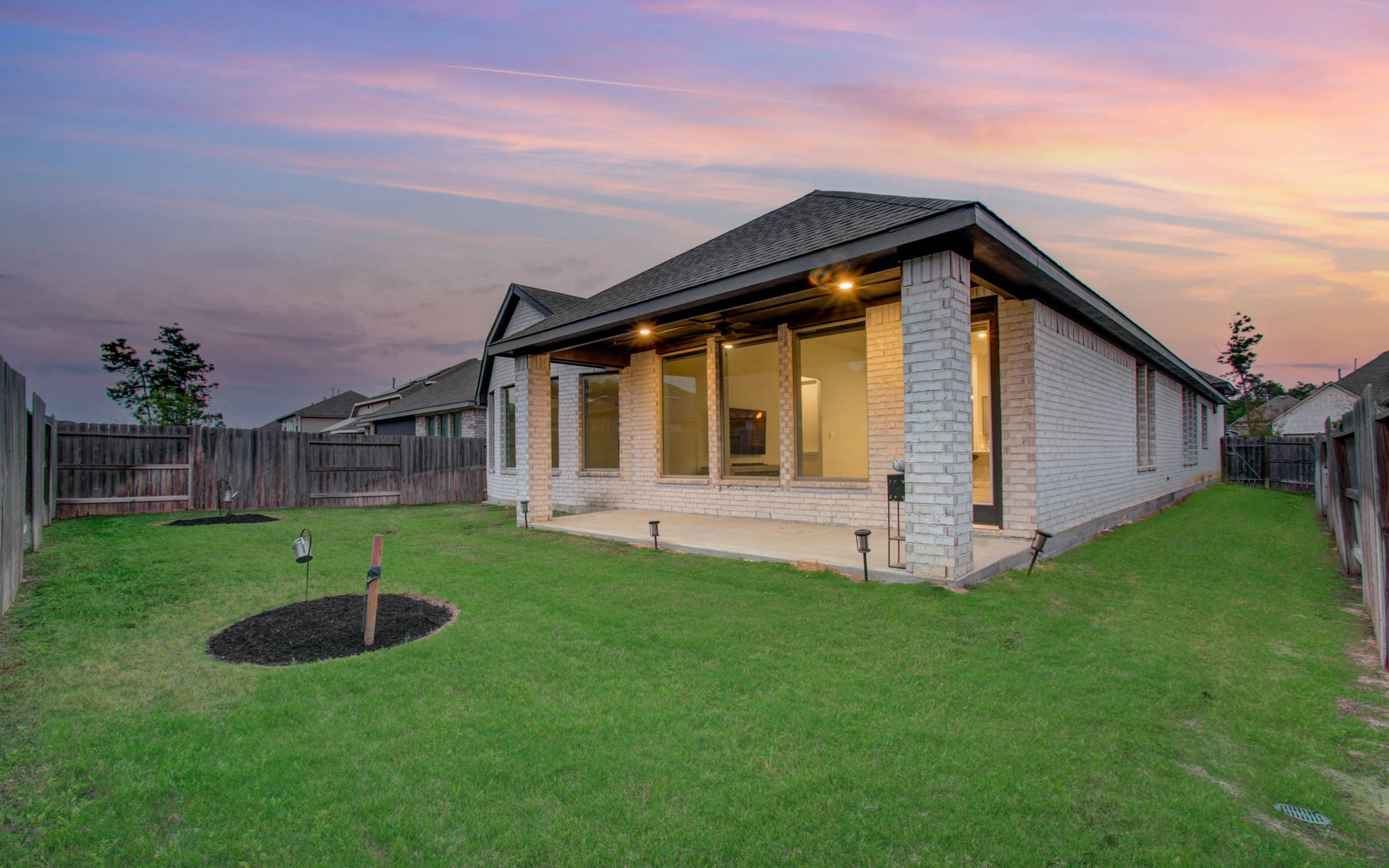 17344 Grand Canyon Road Conroe, TX 77302 - Photo 45 of 49 This photo showcases the backyard of a home at sunset, featuring a covered patio area with a lighted ceiling, brick exterior, and a fenced grassy yard with a small landscaped area.