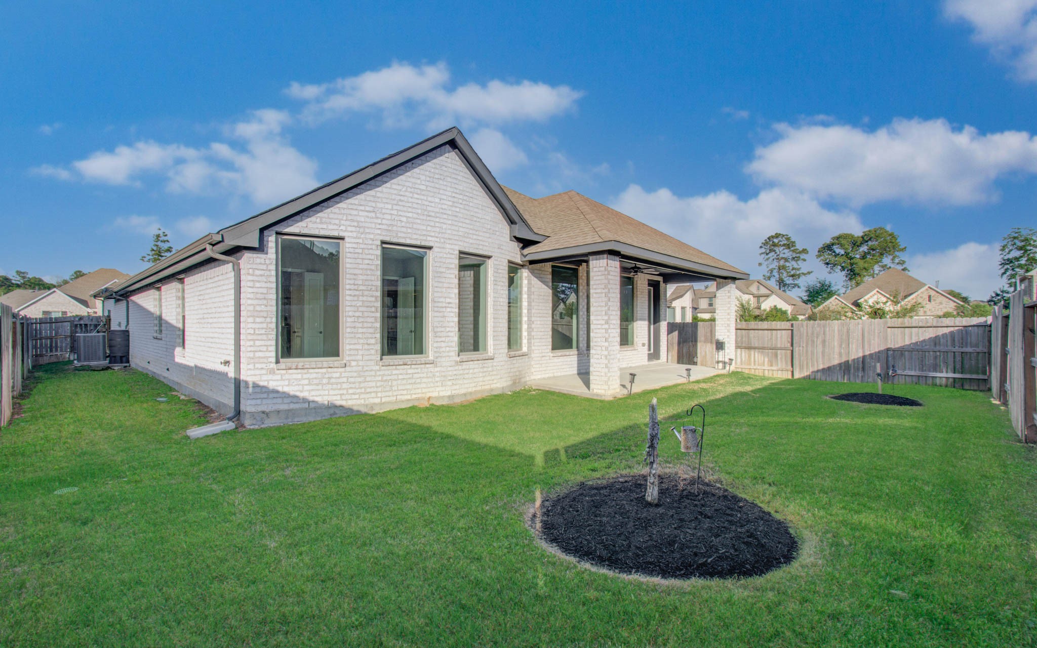17344 Grand Canyon Road Conroe, TX 77302 - Photo 49 of 49 This photo shows a modern single-story white brick house with large windows and a fenced backyard. The yard is neatly landscaped with a small patio area, and the neighborhood appears quiet and residential.