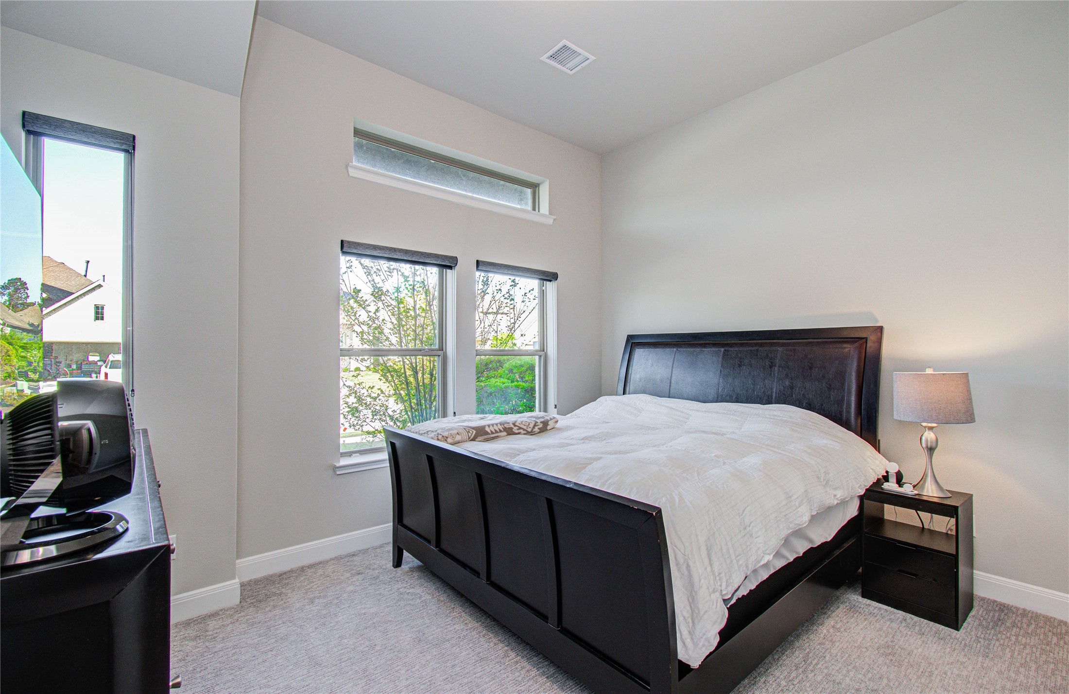 17344 Grand Canyon Road Conroe, TX 77302 - Photo 5 of 49 Bright and inviting bedroom with large windows, neutral walls, and carpeted flooring. It features a spacious bed with a sleek dark frame, matching nightstand, and modern lamp, creating a cozy and elegant atmosphere.