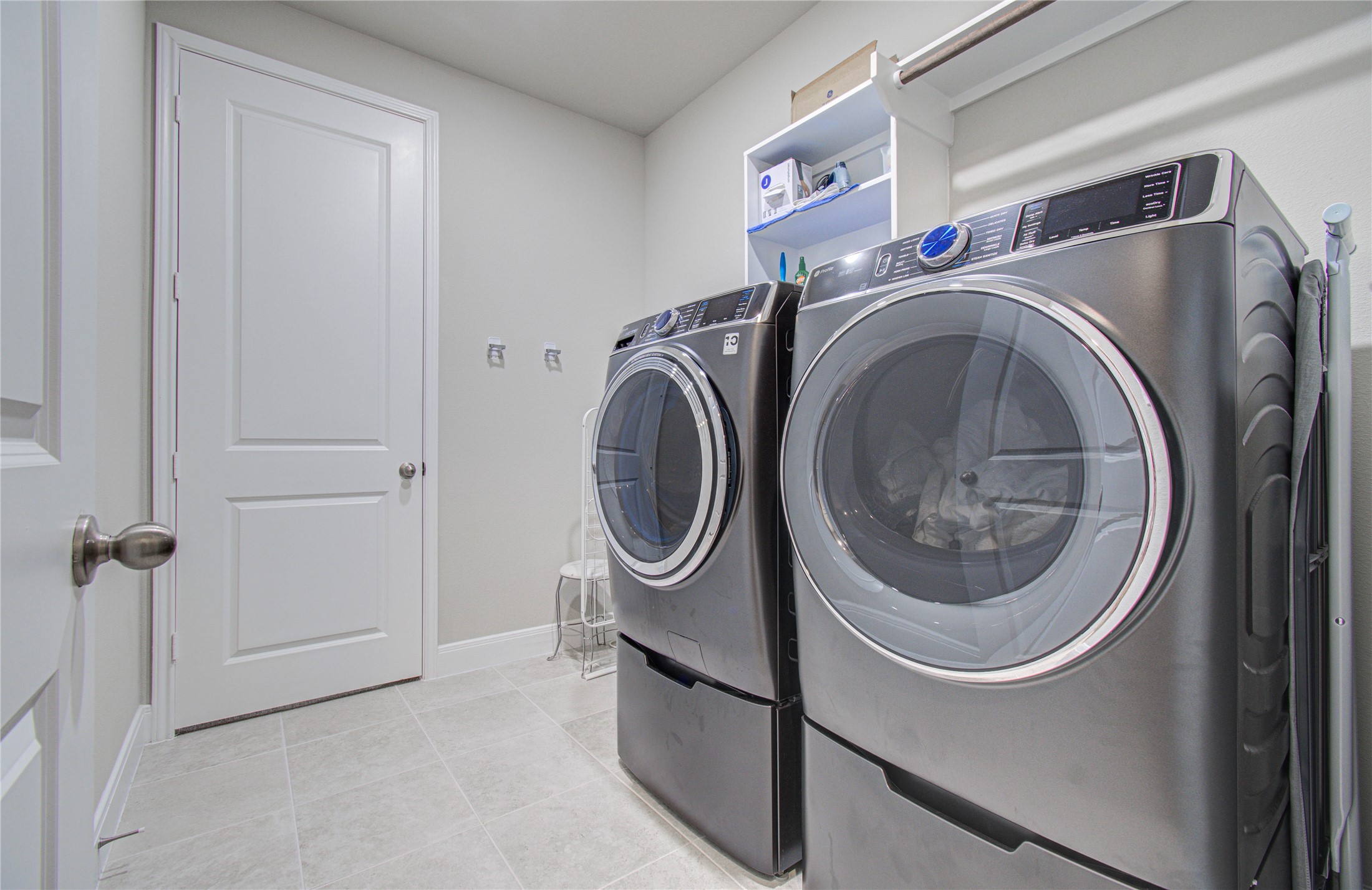 17344 Grand Canyon Road Conroe, TX 77302 - Photo 10 of 49 This photo showcases a modern laundry room featuring a sleek washer and dryer set. It has light-colored tile flooring, wall-mounted shelving for storage, and a closed door. The space is well-organized and functional.