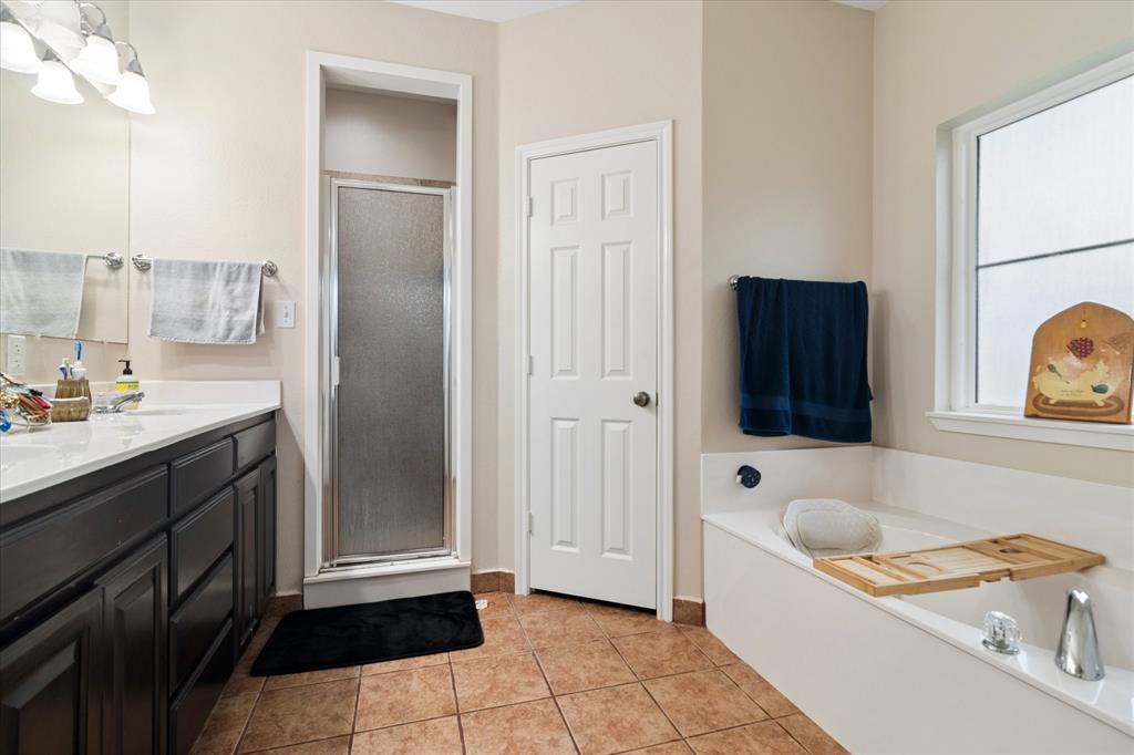 6005 Hawkeye Road Krum, TX 76249 - Photo 17 of 27 Spacious bathroom featuring a white soaking tub, a stand-up shower with frosted glass, a vanity with dark cabinetry and white countertop, a multi-light fixture, and ceramic tile flooring