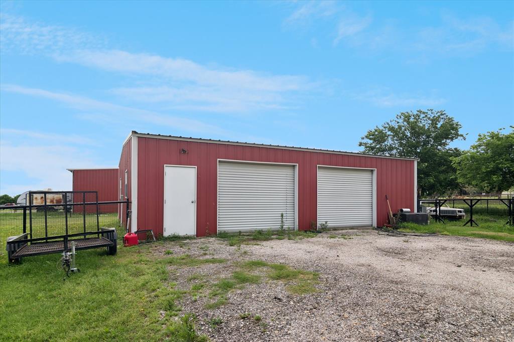 6005 Hawkeye Road Krum, TX 76249 - Photo 24 of 27 Red metal-sided outbuilding featuring two roll-up garage doors, a pedestrian entrance, and a gravel approach