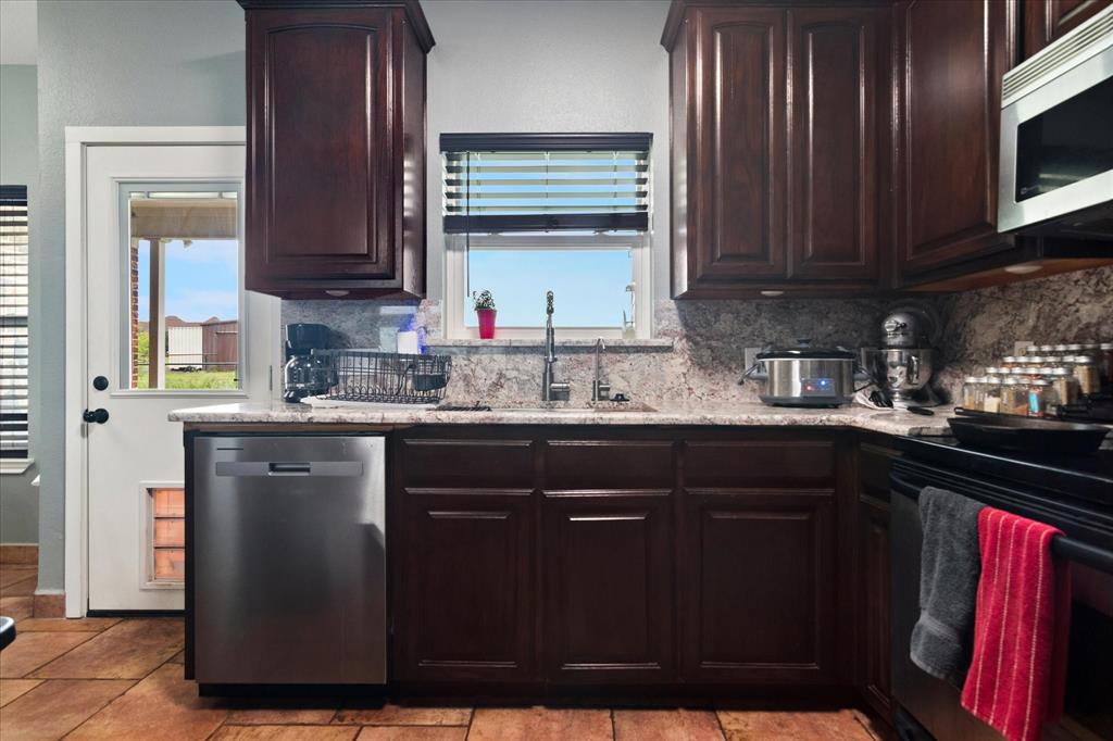 6005 Hawkeye Road Krum, TX 76249 - Photo 4 of 27 Kitchen featuring dark wood cabinetry, a stainless steel dishwasher, a window with horizontal blinds, a stainless steel sink with a high-arc faucet, and granite countertops with a matching backsplash