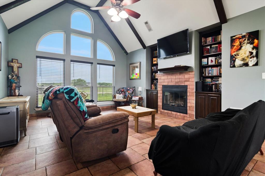 6005 Hawkeye Road Krum, TX 76249 - Photo 9 of 27 Vaulted ceiling featuring exposed beams and a ceiling fan, large arched windows, brick fireplace with mantel, built-in bookshelves, and tile flooring