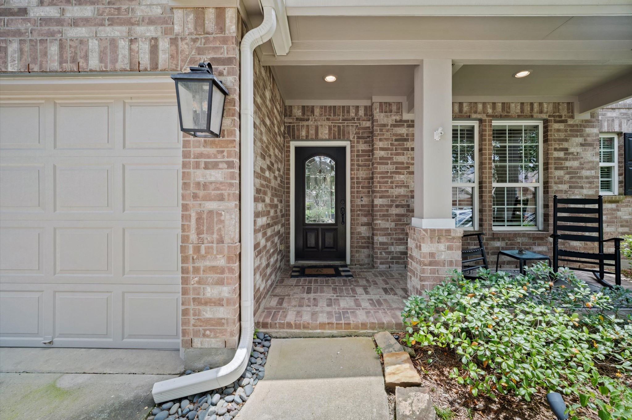 9507 Empress Crossing Drive Spring, TX 77379 - Photo 5 of 50 This inviting home features a charming brick exterior with a cozy covered porch. The entryway is framed by a classic black door with glass detailing. Adjacent is a three-car garage, and the porch area includes seating, perfect for relaxing. Lush landscaping surrounds the entrance, adding to the welcoming feel.