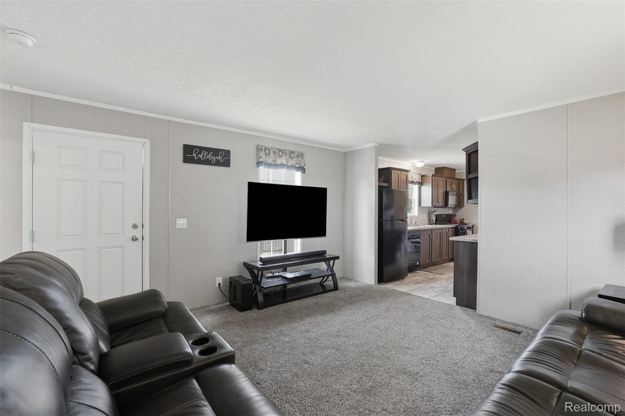 92 Normandy Drive Howell, MI 48843 - Photo 5 of 24 Living area featuring gray wall panels, carpet flooring, a white paneled door, and a partially visible kitchen with dark wood-finish cabinetry