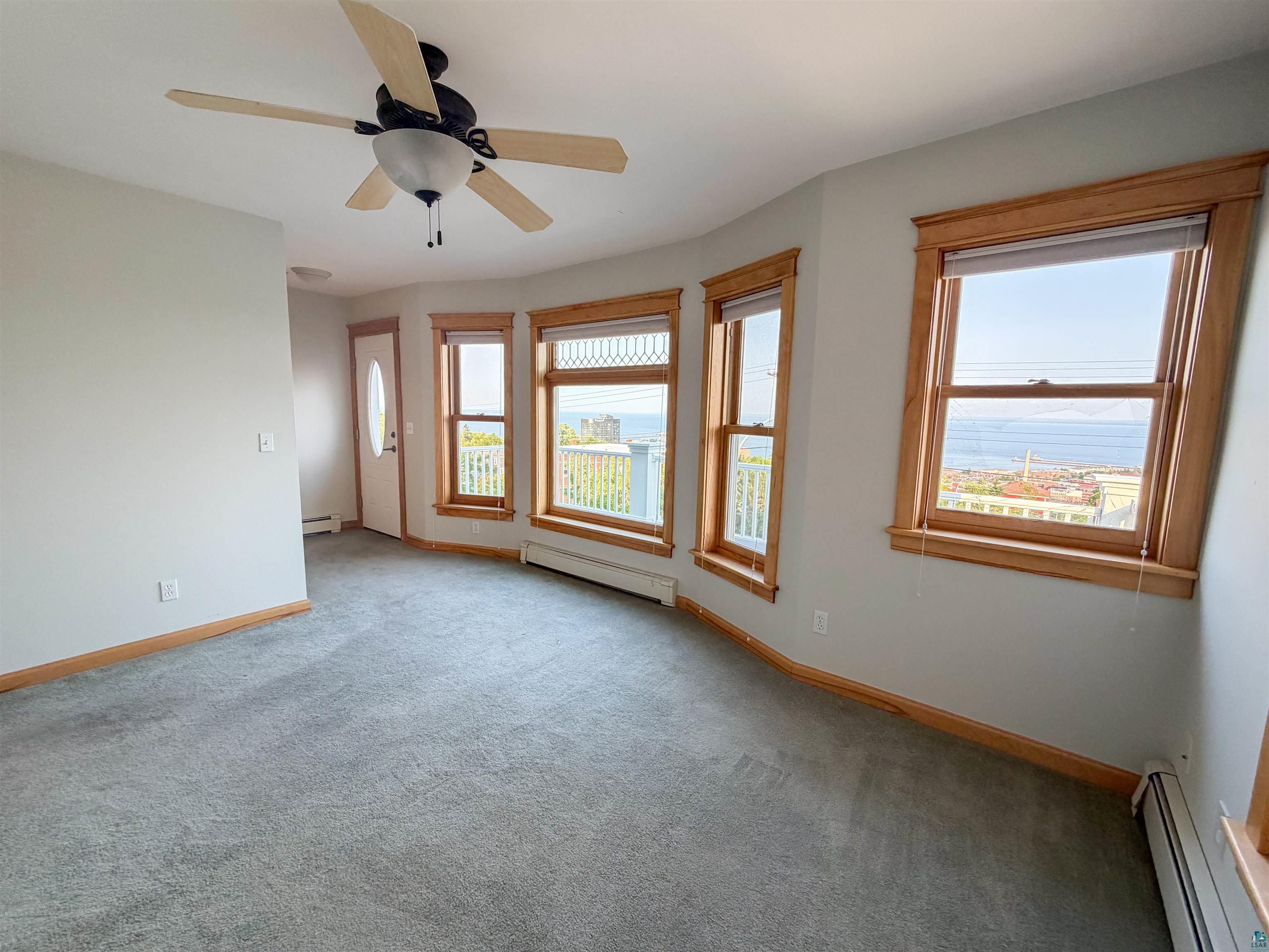 13 West 8th Street Duluth, MN 55806 - Photo 26 of 57 Living area featuring light gray wall paint, gray carpet flooring, wood-finish window trim, a ceiling fan with light fixture, and a bay window configuration with a distant water scene