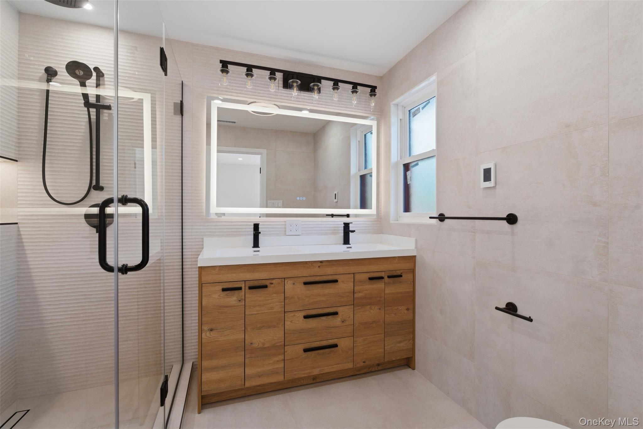 40 Calhoun Avenue New Rochelle, NY 10801 - Photo 23 of 49 Bathroom featuring a double vanity with a light wood-finish cabinet and white countertop, an illuminated mirror, matte black fixtures, and a glass-enclosed shower with a linear drain and heated floor including inside the shower.
