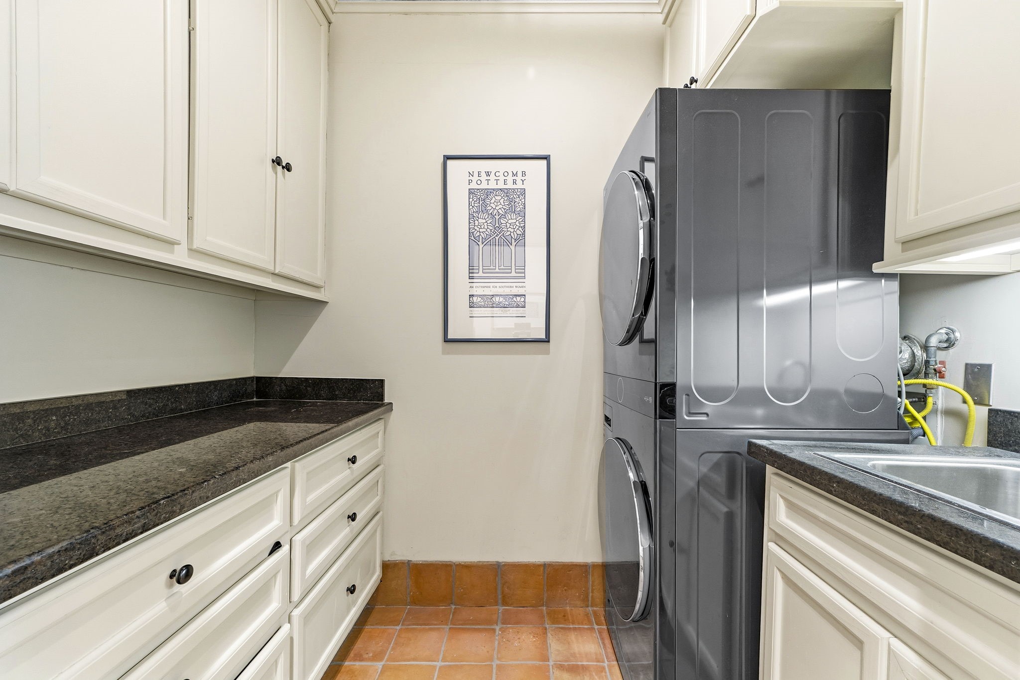 1521 Kipling Street Houston, TX 77006 - Photo 19 of 35 This photo showcases a compact laundry room with stacked washer and dryer units. It features ample storage with white cabinetry and dark countertops, alongside a functional sink area. The space is completed with terracotta floor tiles and a framed wall art piece.