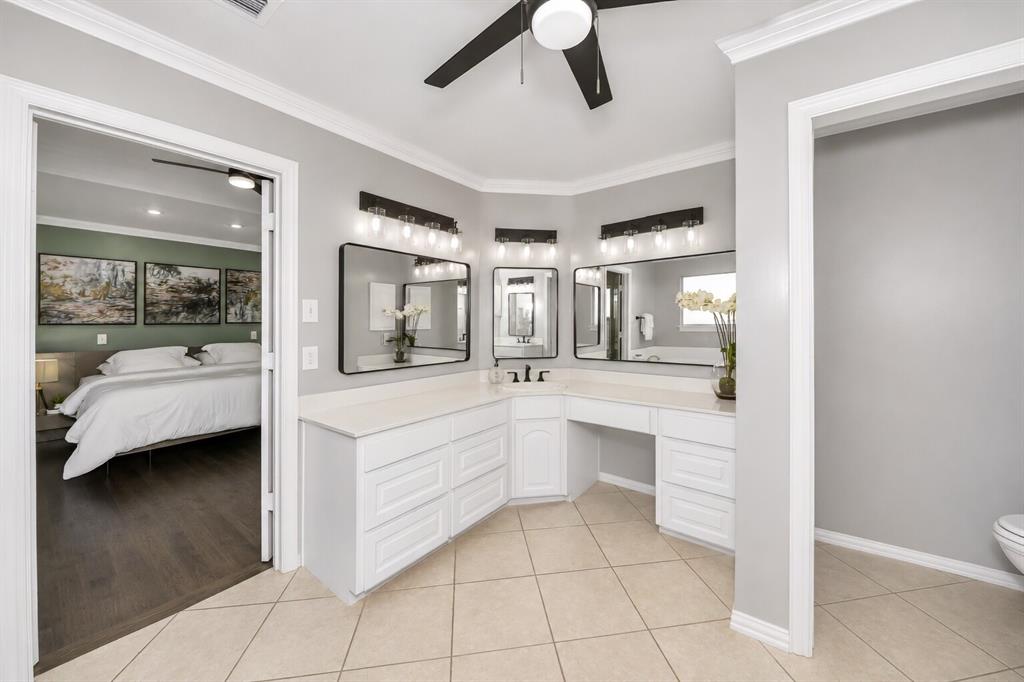 3817 Ganzer Road West Denton, TX 76207 - Photo 28 of 40 The primary bathroom features a corner three-station vanity with space for a chair, designed for comfortable everyday routines. Framed mirrors and soft, flattering lighting create a bright, modern look that keeps the space feeling calm and functional. White cabinetry and black accents tie everything together, giving the room a timeless style that’s easy to live with.