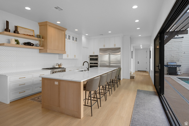 5367 Cyan Lane St. George, UT 84790 - Photo 26 of 49 Gourmet kitchen featuring a large central island with seating, stainless steel appliances, white cabinetry with gold-tone hardware, a wood-finish range hood, and light wood-finish flooring