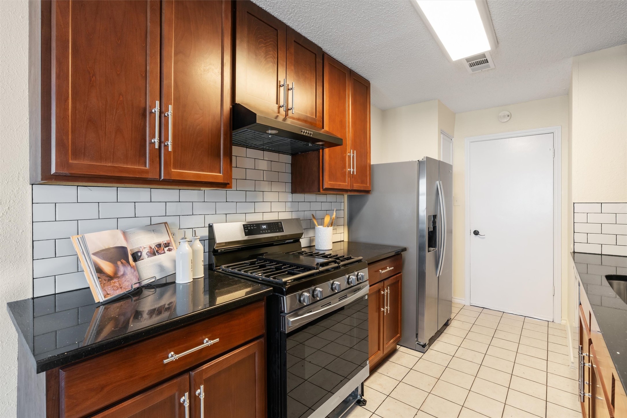 11902 Barrington Way Austin, TX 78759 - Photo 10 of 30 Kitchen featuring stainless steel appliances, a textured ceiling, light tile patterned flooring, wood finish cabinetry, and dark stone counters