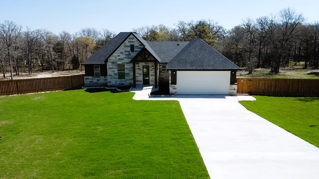 View of front of home with stone siding, driveway, a garage, view of wooded area, and a shingled roof