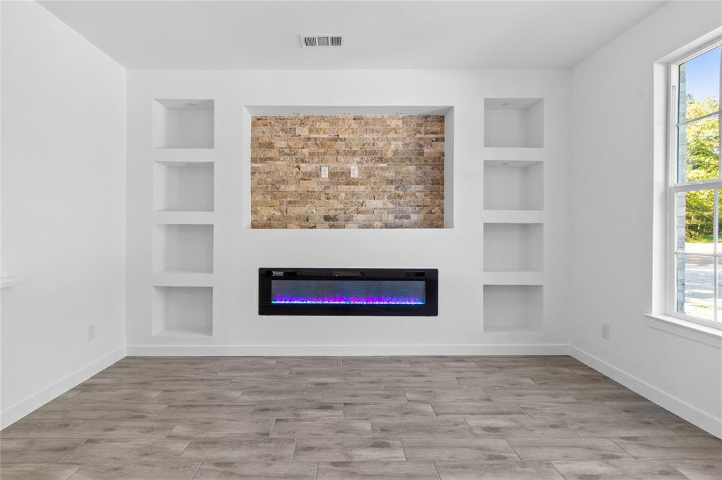 7520 Double Bridge Road Mabank, TX 75156 - Photo 7 of 32 Modern entertainment space featuring custom built-in shelving flanking a brick accent wall, linear electric fireplace with blue LED accent lighting, wood-finish plank flooring, and abundant natural light from large picture window