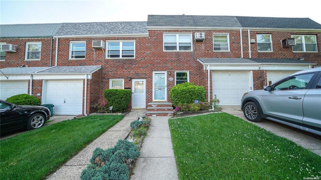 View of property featuring driveway, a front yard, a wall mounted air conditioner, a garage, and brick siding