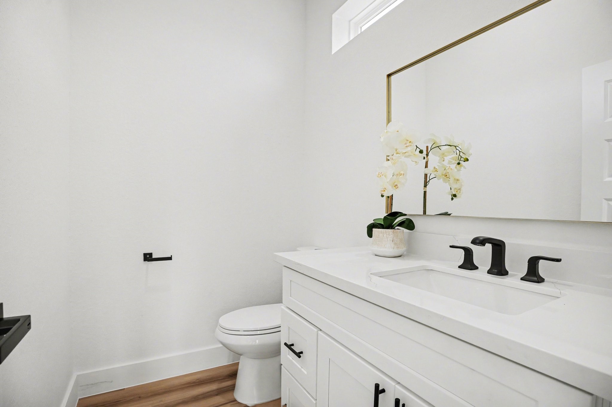 115 Milby Street Houston, TX 77003 - Photo 12 of 28 This photo showcases a modern and bright half bathroom with white walls and wood flooring. It features a sleek white vanity with black fixtures, a large mirror, and a potted plant for decoration.