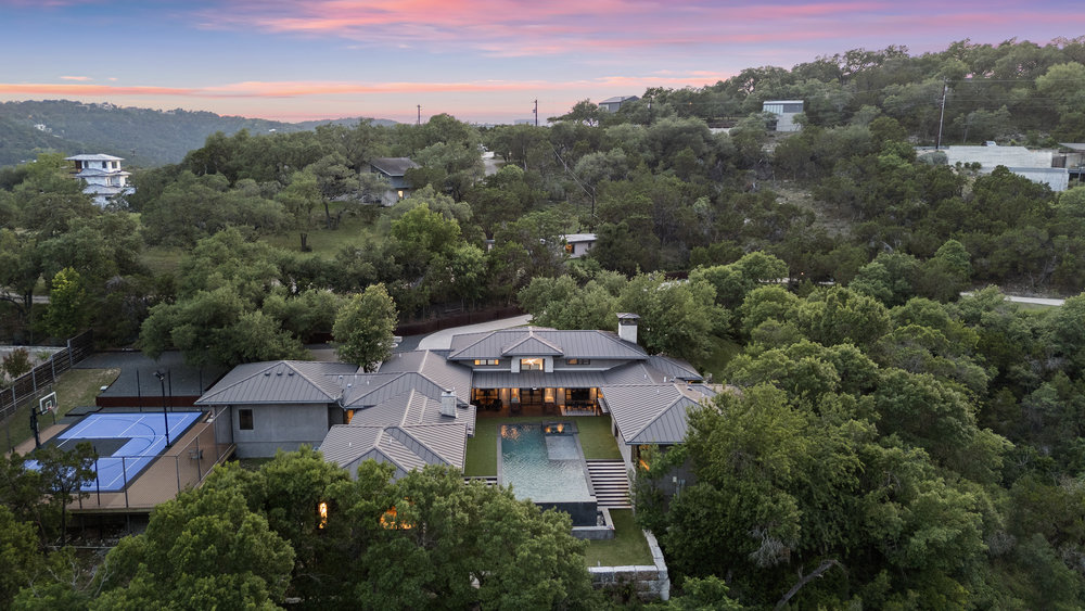 1905 Paisano Road Austin, TX 78746 - Photo 38 of 40 An aerial panorama reveals a sprawling contemporary residence nestled among verdant hills, featuring a private pool and sport court.