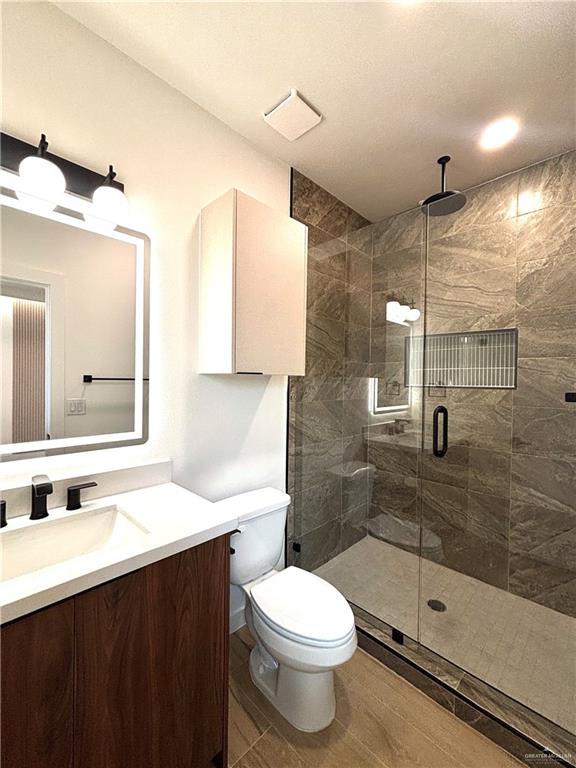 4 Estevan Street Mission, TX 78573 - Photo 23 of 24 Modern bathroom featuring a wood-finish vanity with a white countertop and rectangular sink, matte black fixtures, an illuminated mirror, and a glass-enclosed shower with stone-look wall tiles and a rainfall showerhead