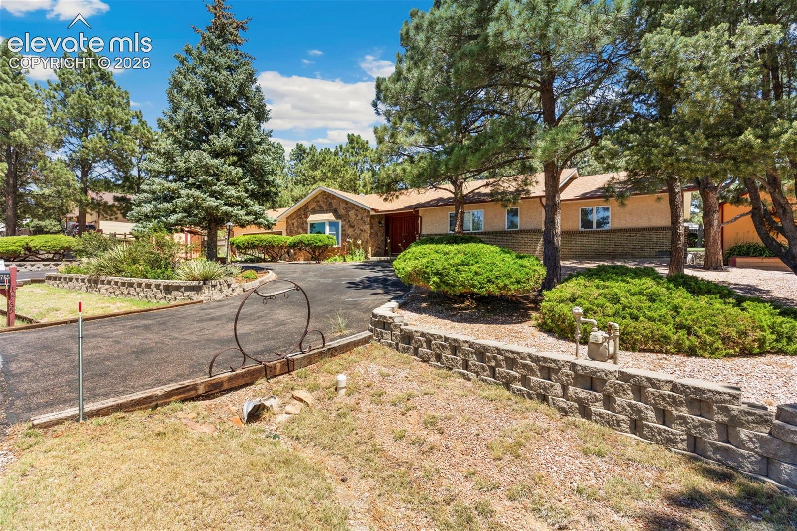 10765 Falling Star Road Fountain, CO 80817 - Photo 2 of 27 Ranch-style residence featuring a stone and stucco exterior, mature trees, a paved driveway, and tiered landscaping with retaining walls