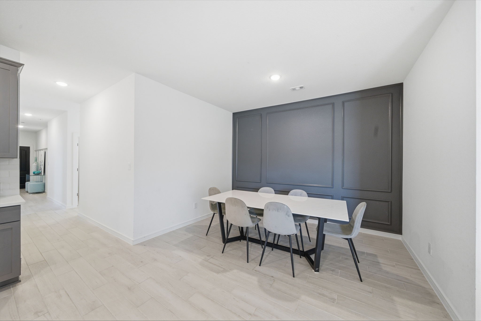 302 Alder Loop Anahuac, TX 77514 - Photo 29 of 34 This photo showcases a modern dining area with a sleek white table and six chairs. It features a dark accent wall, light wood flooring, and an open hallway leading to other spaces.