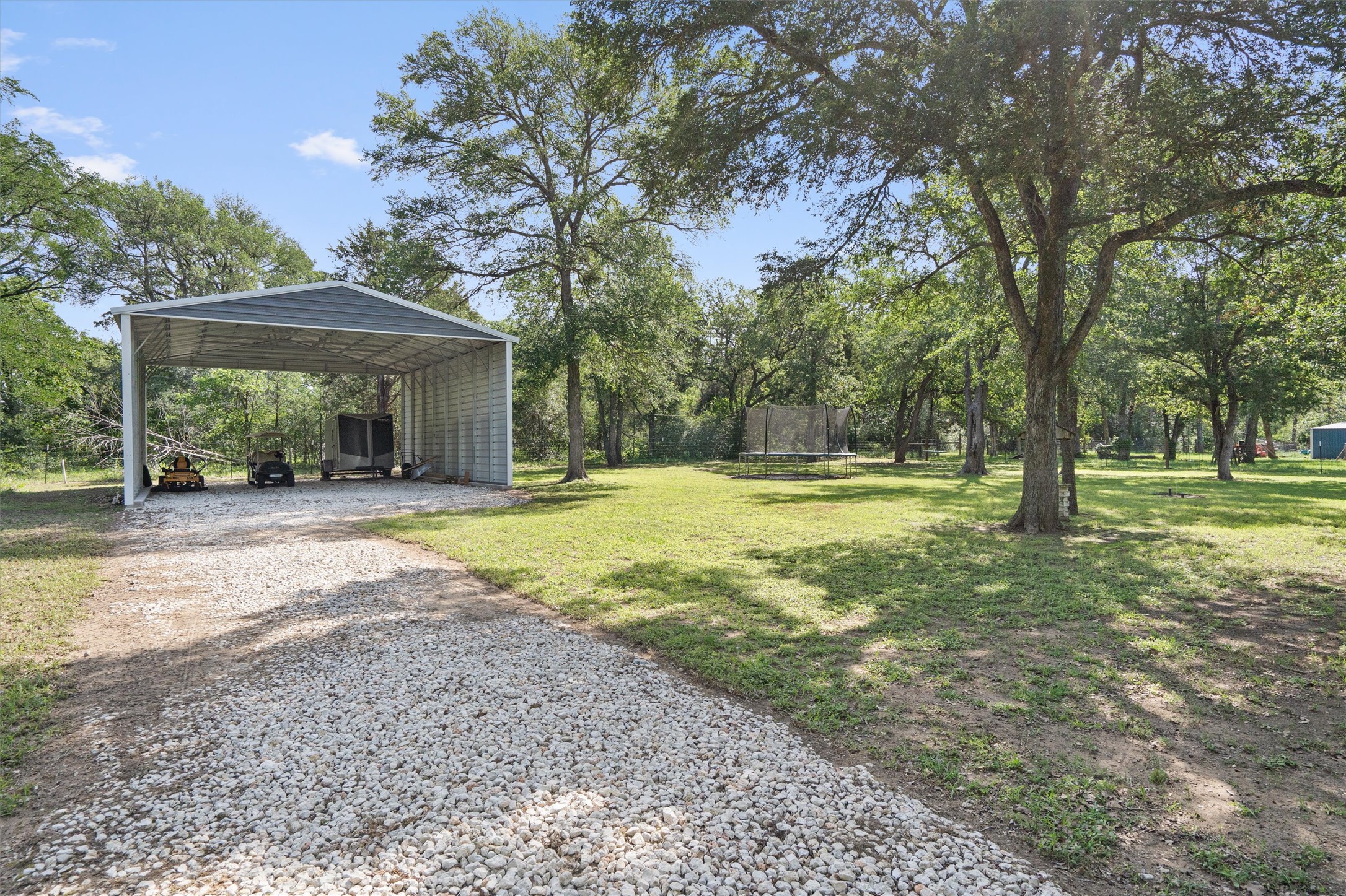 136 Fawn Hollow Elgin, TX 78621 - Photo 38 of 40 Gravel driveway leading to a metal carport structure with an expansive, treed lot featuring mature trees and a grassy expanse