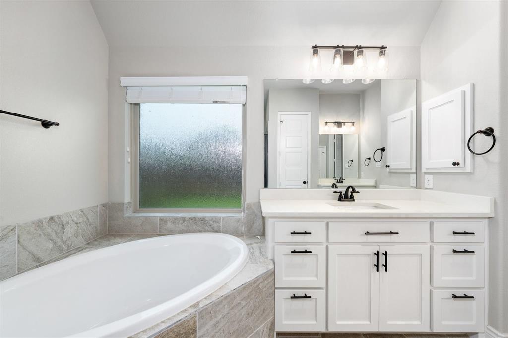 1501 Calcot Lane Forney, TX 75126 - Photo 19 of 34 Soaking tub with a tiled surround, frosted window, and a white vanity featuring a single undermount sink, white countertop, and black hardware