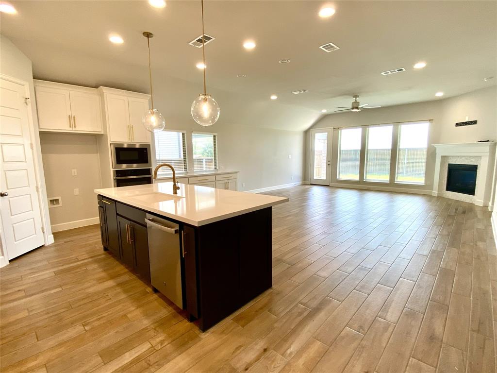 1937 Halkin Road Celina, TX 75009 - Photo 14 of 20 Open-concept space featuring a kitchen island with sink and stainless steel dishwasher, wood-like tile flooring, white cabinetry, a built-in microwave and oven, and a fireplace with a white mantel