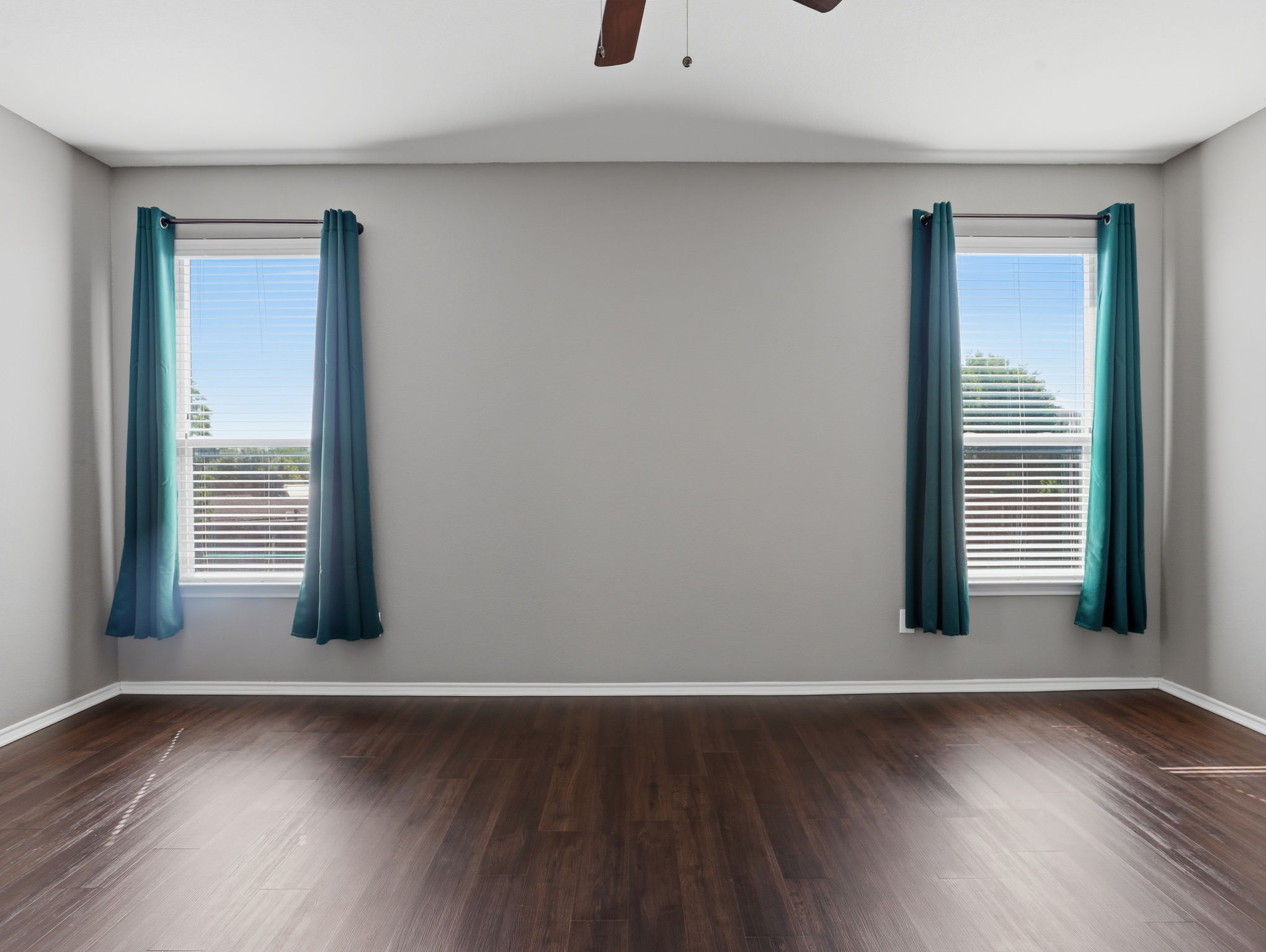 640 Landon Samuel Loop Pflugerville, TX 78660 - Photo 20 of 33 Spacious room featuring wood-finish flooring, light gray walls, two windows with blinds and teal curtains, and a ceiling fan